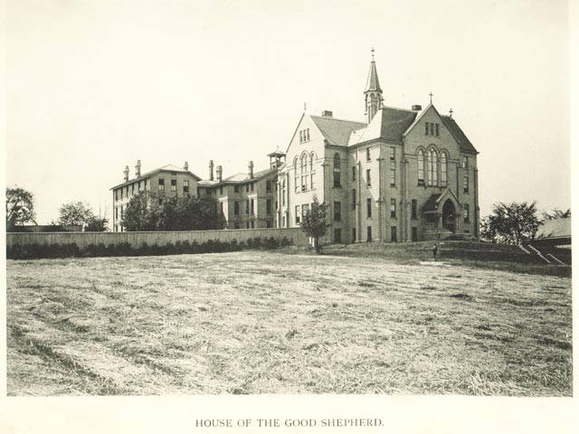 A black-and-white photograph of a large brick building on top of a hill. A empxty field is front of the building, and a fence extends from the building.